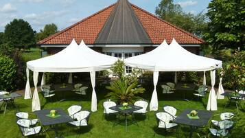 Ein Außenbereich mit Terrassenmöbeln, einer weißen Zelthalle und einem Gebäude mit rotem Dach im Hintergrund.