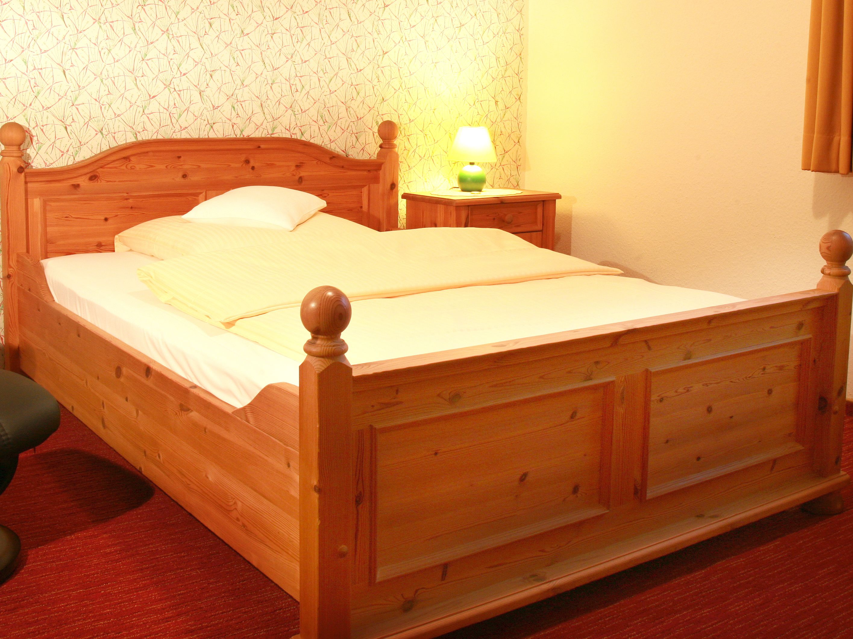 A wooden bed with a headboard, a bedside table with a lamp, and a patterned wallpaper.