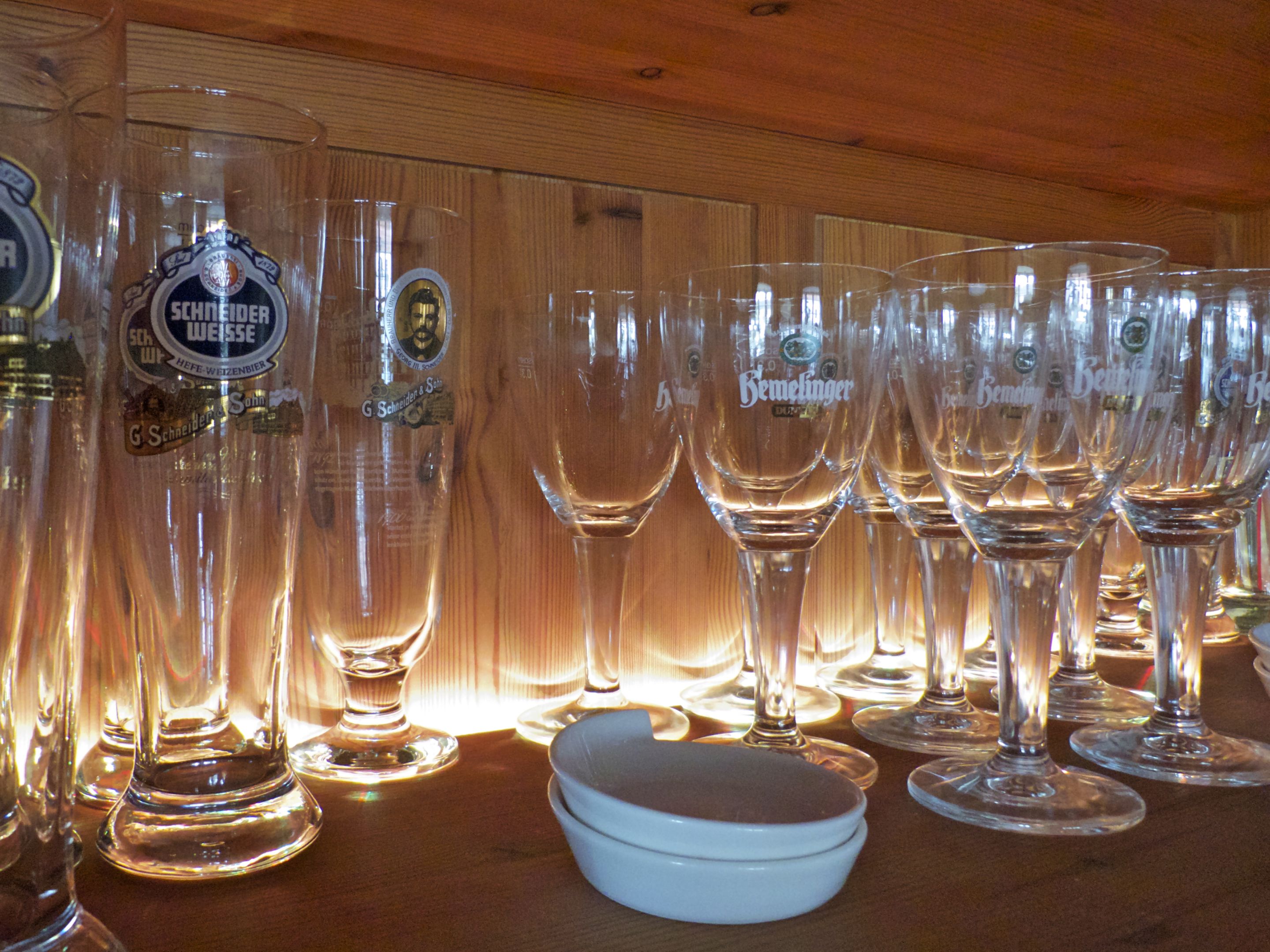 A row of empty wine glasses and a bottle of Schneider Weisse beer on a wooden shelf.