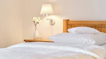 Una cama bien hecha con sábanas blancas, una lámpara de noche y un jarrón con flores blancas sobre una mesita de madera.