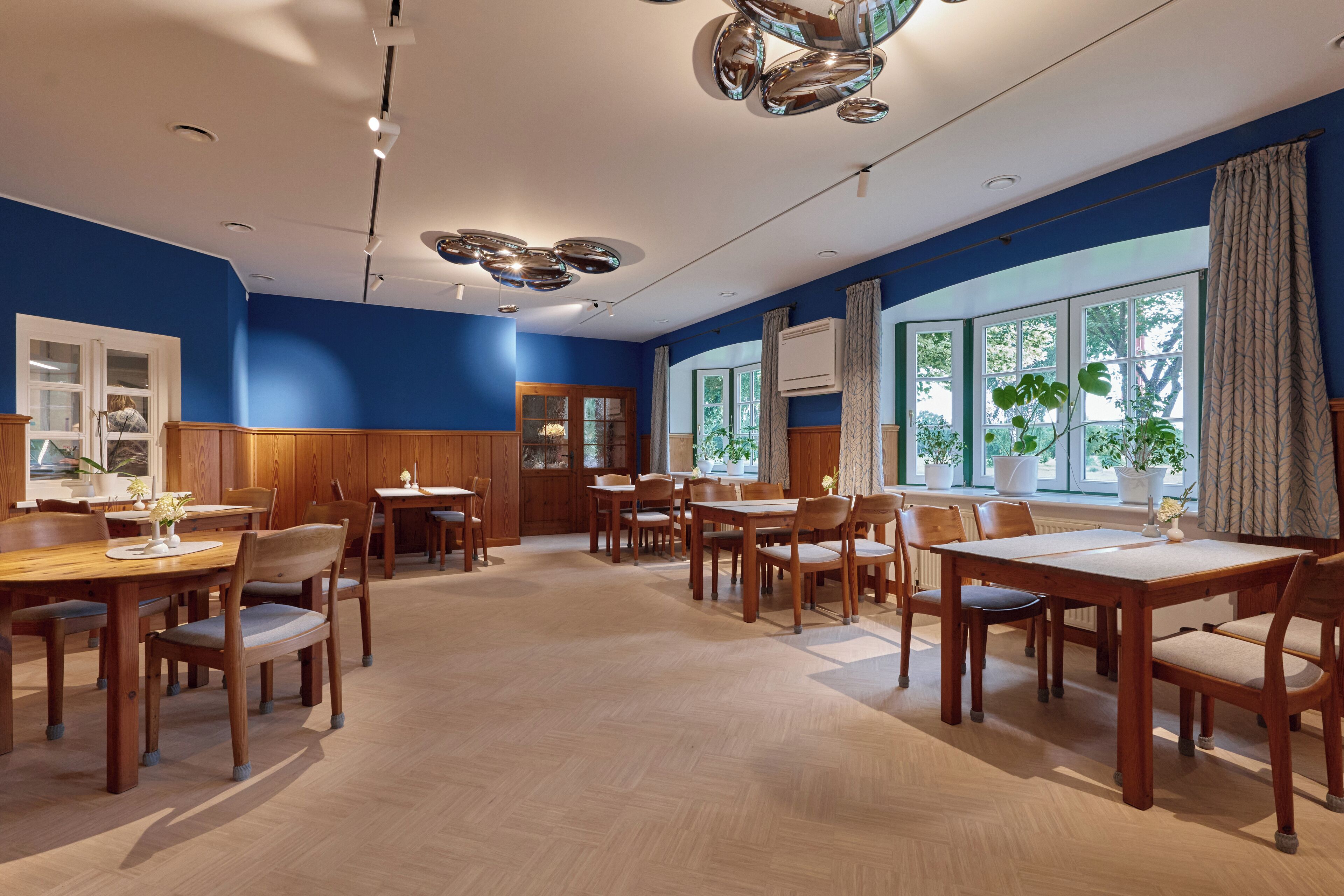 Un comedor amplio con mesas y sillas de madera, ventanas grandes con cortinas y un diseño de techo moderno.