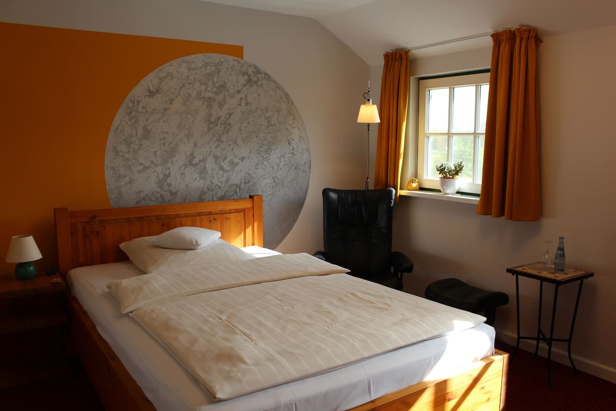 Habitación de hotel con una cama grande, una mesita de noche de madera con lámpara, una silla negra, una pequeña mesa con una botella y vasos, y un ventanal con cortinas.