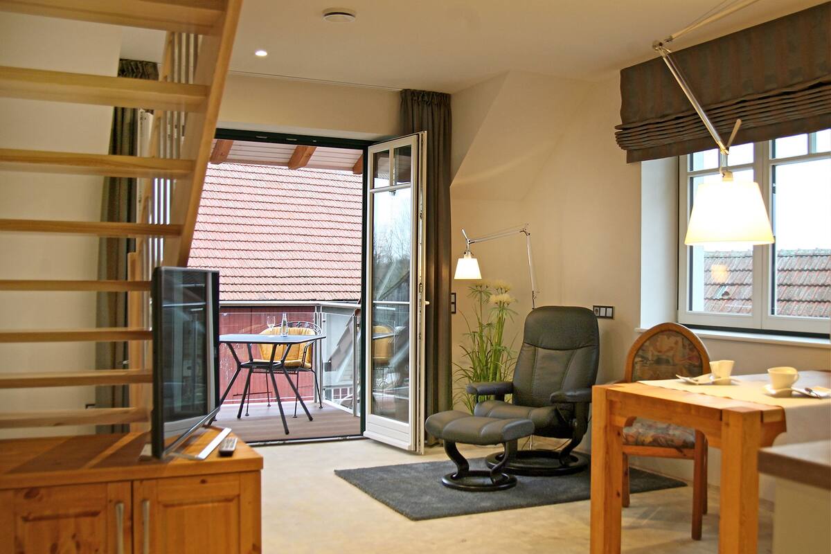 Una habitación con escritorio de madera, una silla, televisor y ventana con vista a un edificio de tejados rojos.