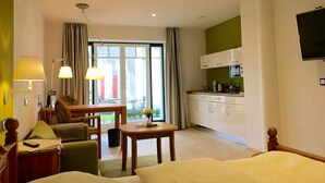 Habitación de hotel con cama, escritorio, silla, mesita, televisor, kitchenette y ventana con cortinas.
