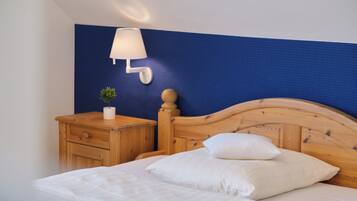 Una cama bien tendida con sábanas y almohadas blancas, una mesita de noche de madera con una lámpara y una pequeña planta en maceta.