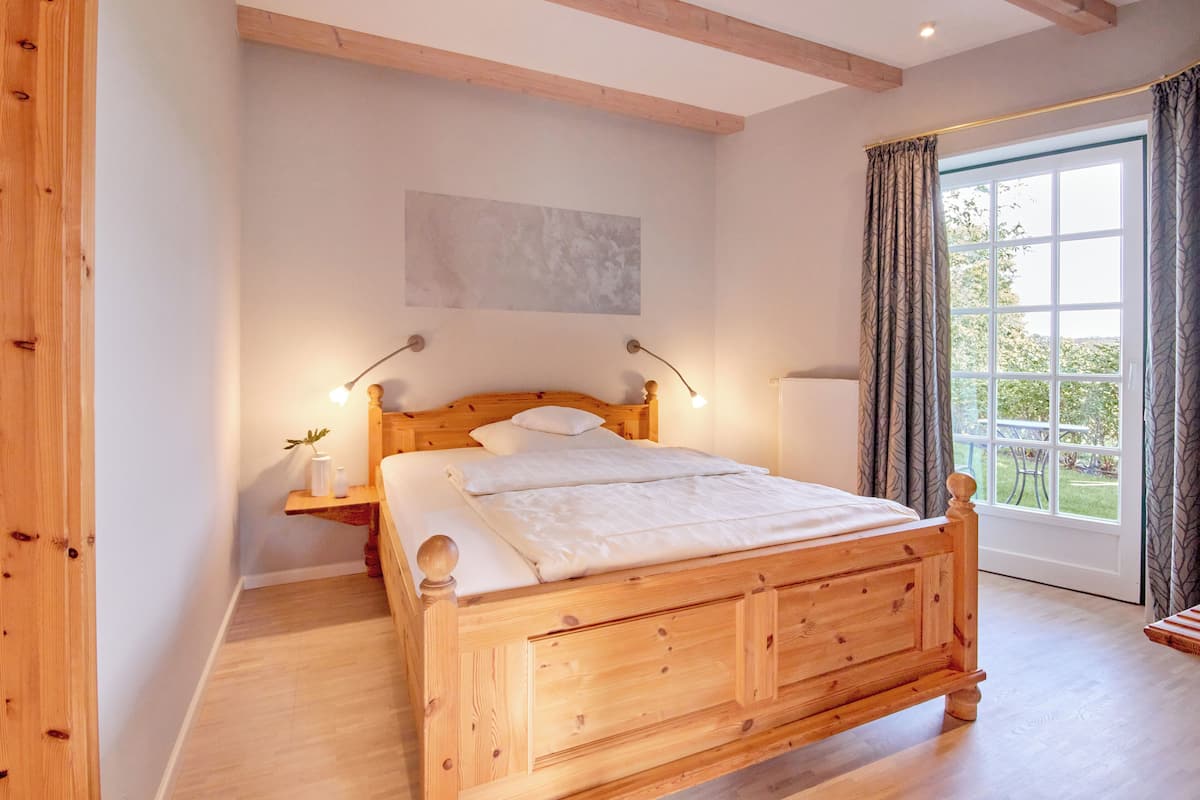Una cama de madera con cabecera, lámparas de noche, una mesita auxiliar con un florero y una ventana con cortinas.