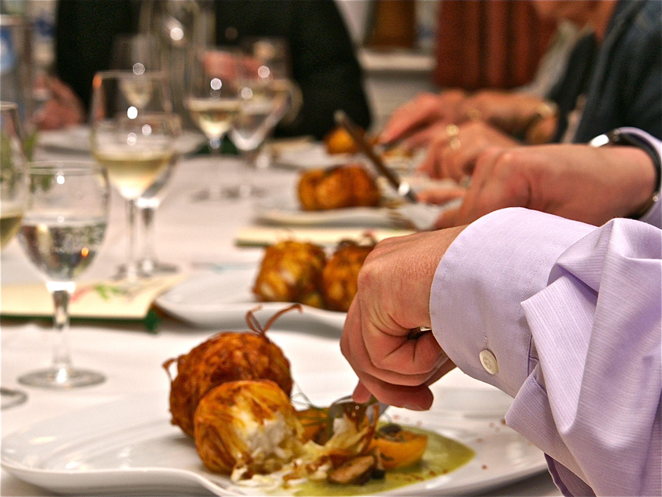 A dining table with plates of food, glasses of white wine, and a person in a purple shirt.