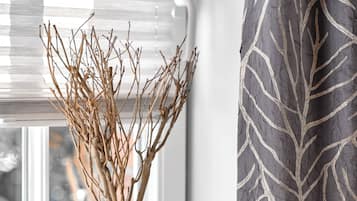A window with a white vase containing branches, a window blind, and a curtain with a leaf pattern.