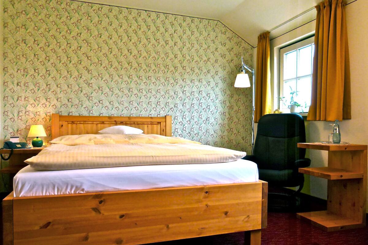 Una cama de madera con ropa de cama blanca, una lámpara de noche, una silla, una mesita de madera y una ventana con cortinas.