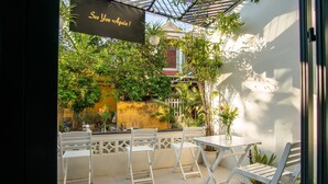 A patio with white chairs and tables, surrounded by lush greenery and a yellow wall. - D Central Homestay Hoi An (Da Nang)