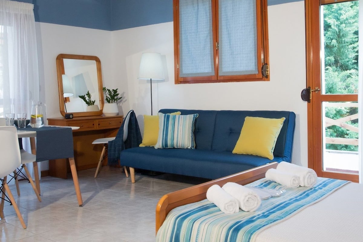 A bedroom with a bed, a desk, a chair, a sofa, a mirror, and a window.