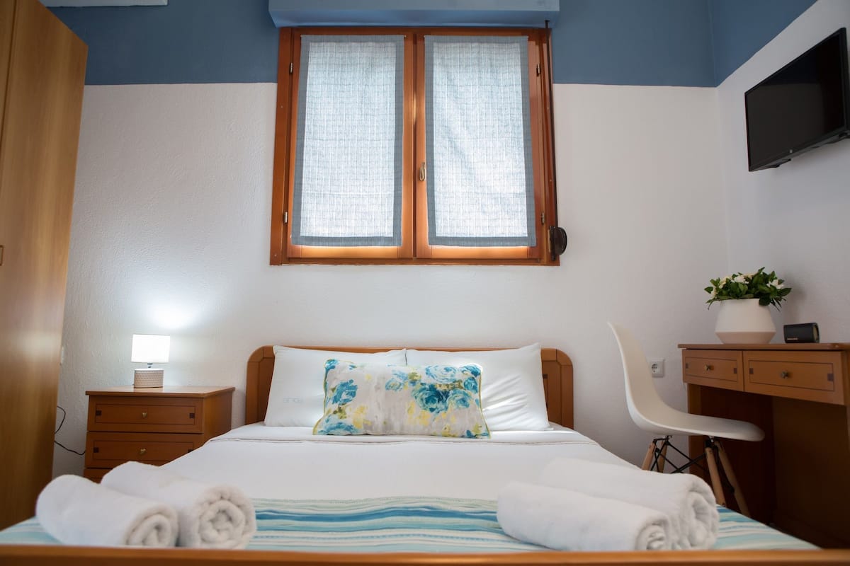 A neatly made bed with a floral pillow, a bedside lamp, a wooden nightstand, a desk with a chair, and a TV mounted on the wall.