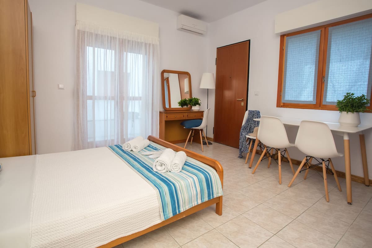 A hotel room with a bed, a desk, two chairs, a window with blinds, and a potted plant.