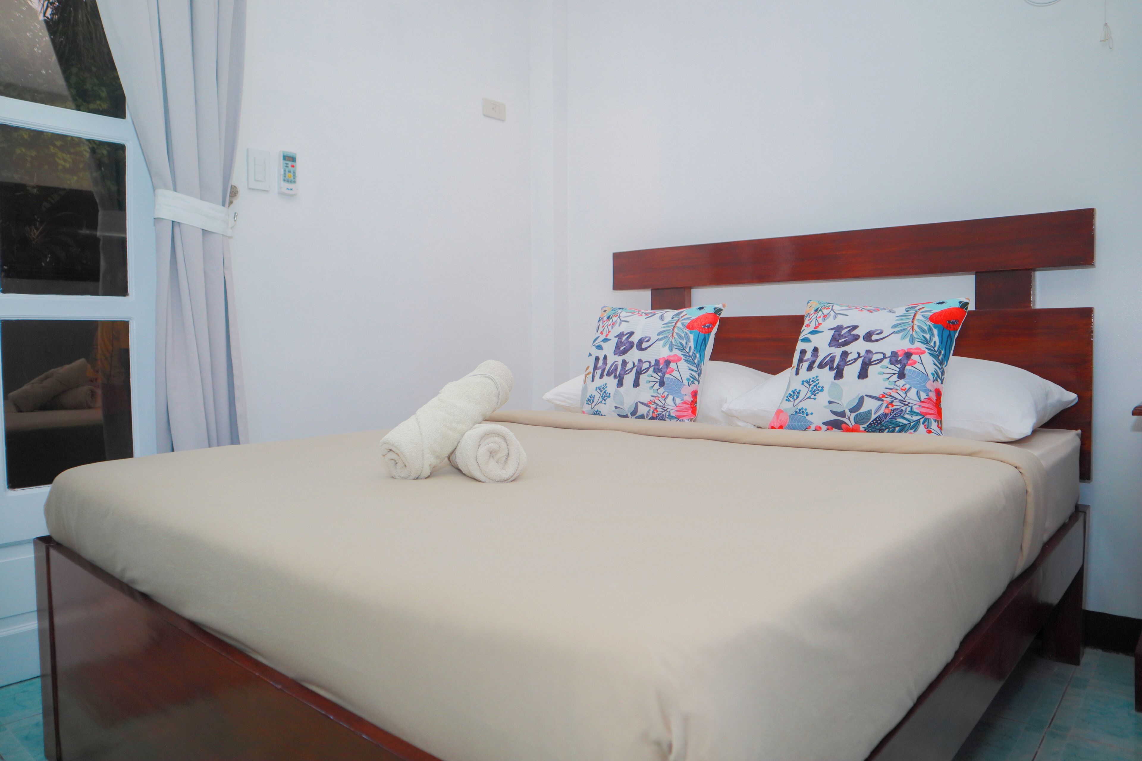 Una cama bien hecha con una colcha color beige, dos almohadillas decorativas con el texto 'Be Happy' y una toalla blanca enrollada.