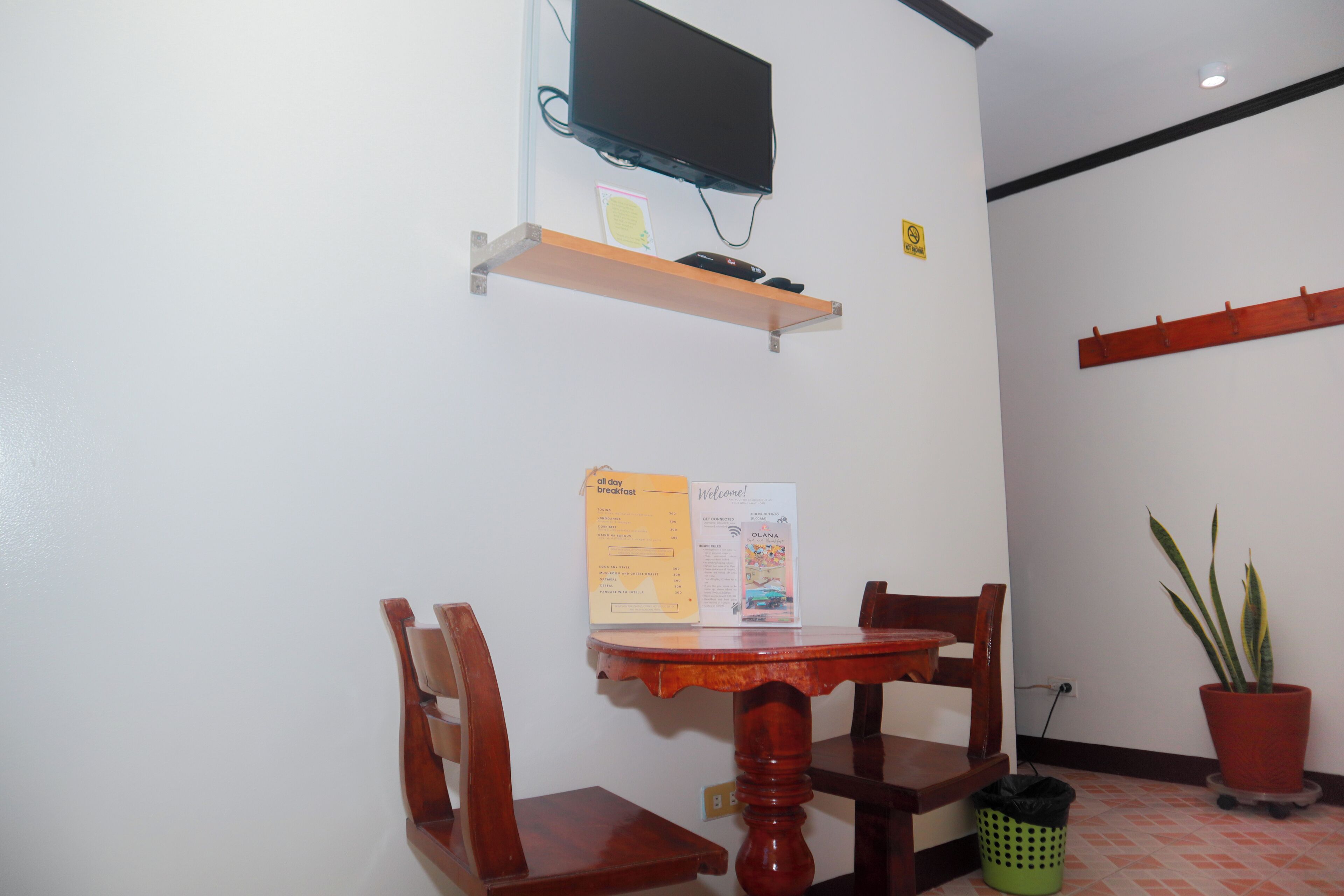 A small dining area with a wooden table and chairs, a television mounted on a shelf, a welcome sign, and a potted plant.