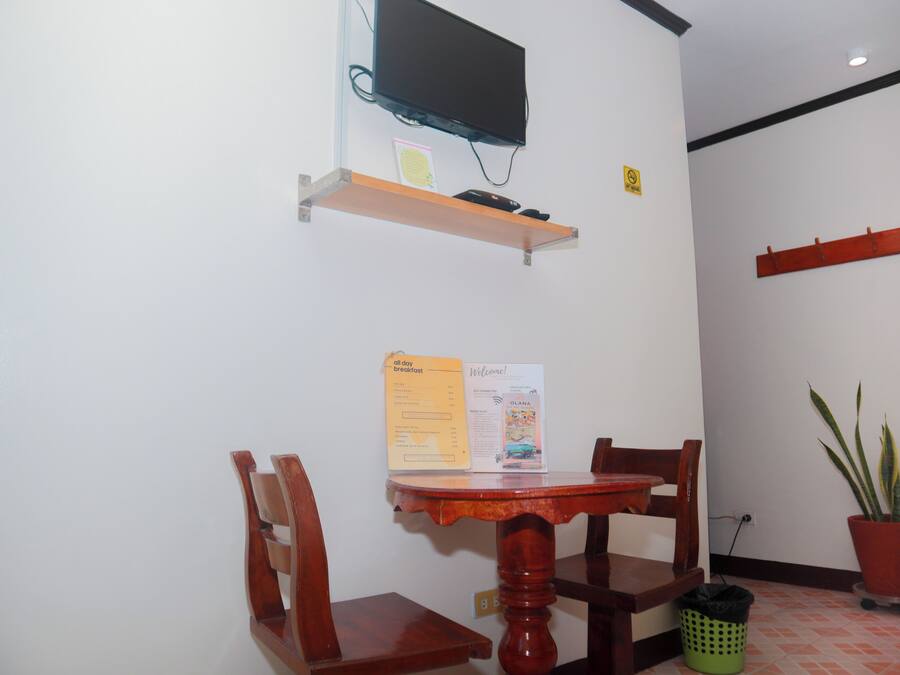 A small dining area with a wooden table and chairs, a television mounted on a shelf, a welcome sign, and a potted plant.