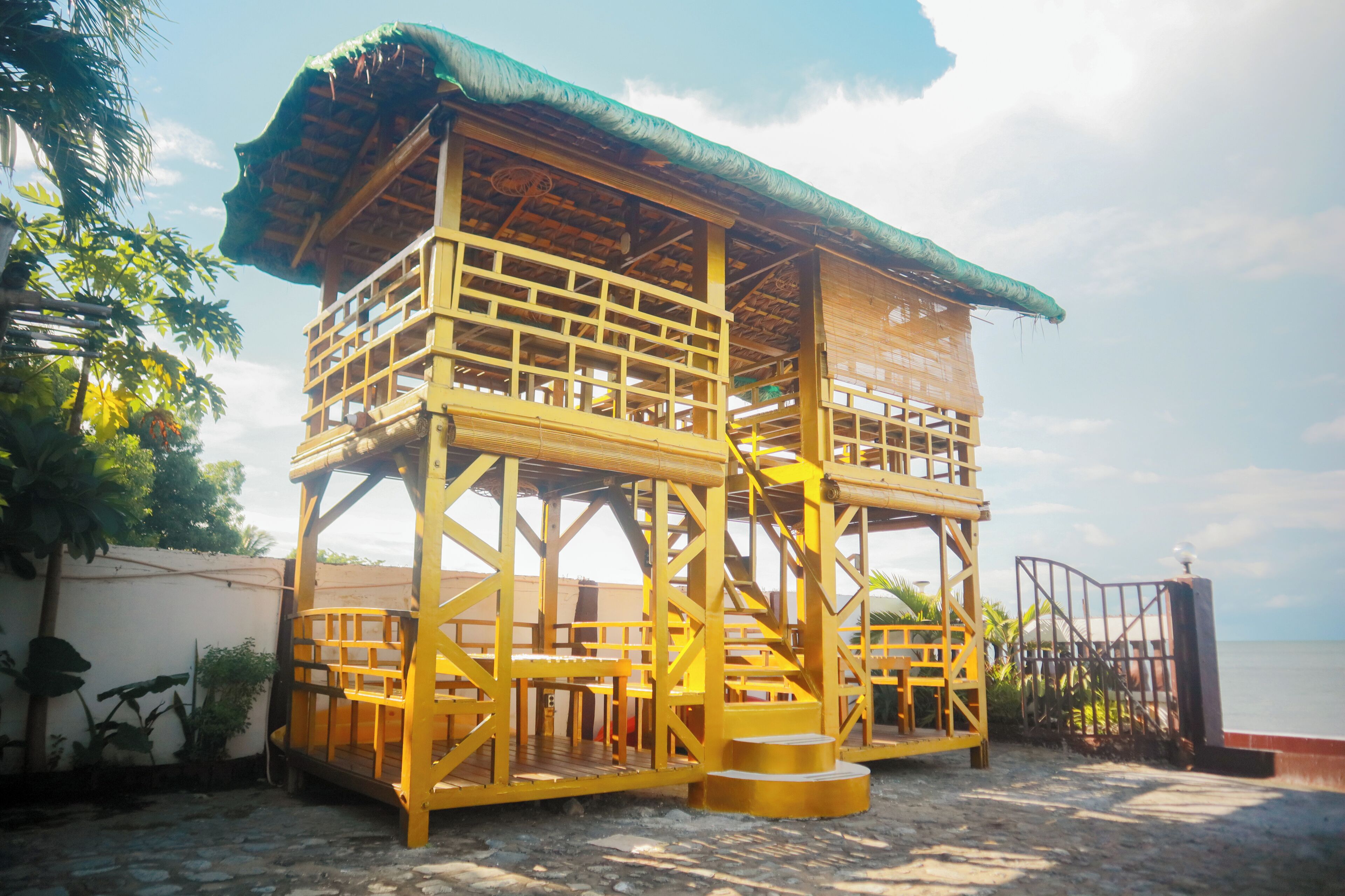 A wooden structure with multiple levels, a staircase, and a thatched roof, situated on a sandy area with a view of the sea.