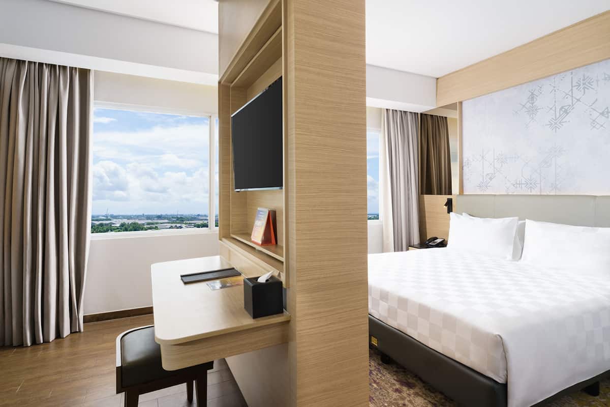 Habitación de hotel con escritorio, cama, ventana grande con cortinas y vistas a la ciudad.