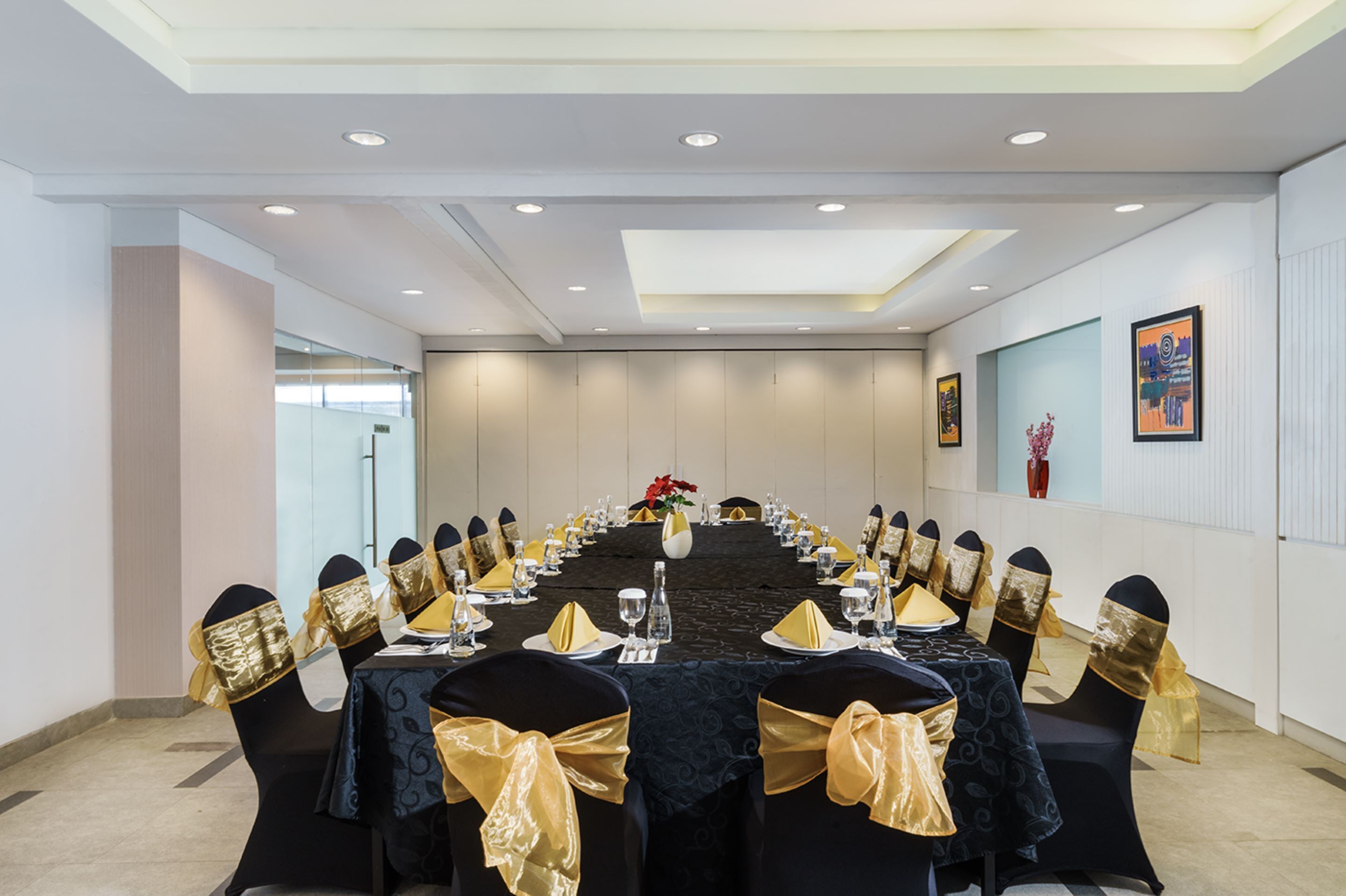 A conference room with a long table set for a meeting, surrounded by chairs with gold sashes, white walls, and ceiling lights.