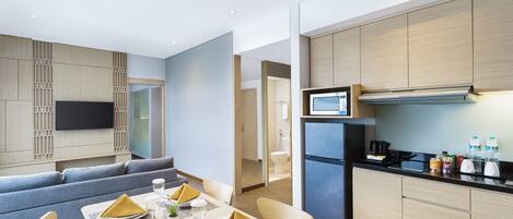 A modern hotel room with a dining area, a sofa, a flat-screen TV, a kitchen with a refrigerator, and a microwave.