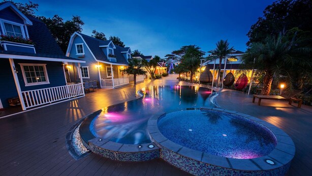 Zona de piscina con jacuzzi, terraza de madera y casas al fondo.