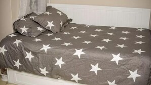 Un lit avec une couvre-lit grise ornée d’étoiles blanches, une tête de lit blanche et une table de chevet blanche avec une lampe.