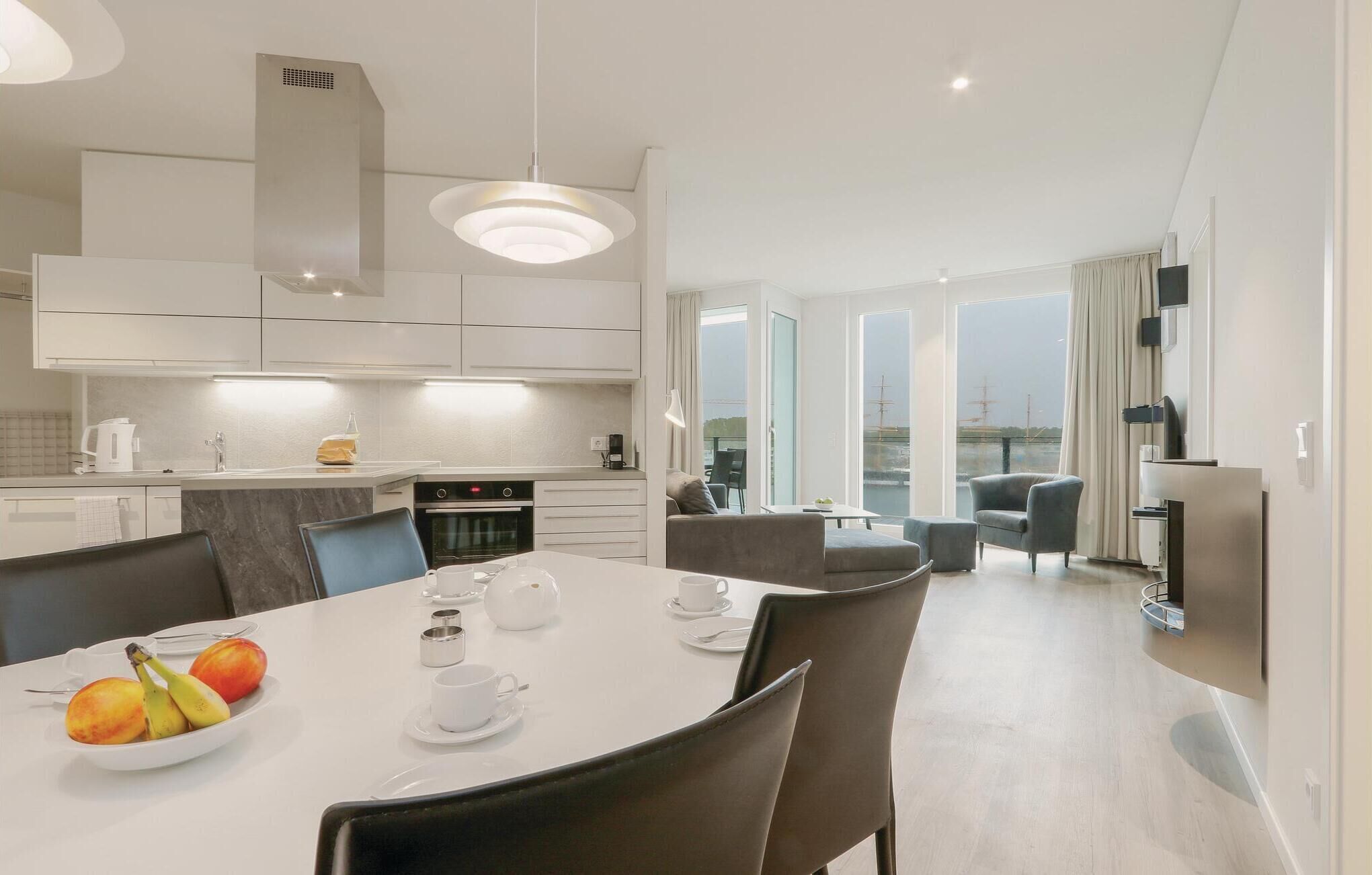 A modern kitchen with white cabinets, a dining area with a white table set for two, and a view of the sea through large windows.