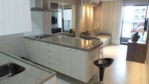 A modern kitchen with a granite countertop, stainless steel sink, and a built-in oven. There is a bar stool near the window, and a sofa in the living area. - Landscape Diamond (Fortaleza)