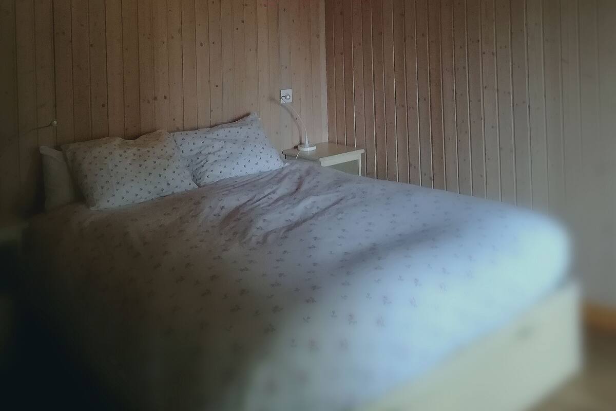 Een bed met een bloemenprint beddekleed en kussens, een nachtkastje met een lamp, en een houten wand.