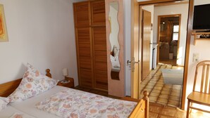Quarto com guarda-roupa de madeira, cama com roupa de cama florida, mesa de cabeceira com abajur e televisão sobre suporte.