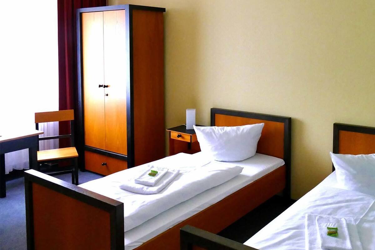 Una habitació d'hotel amb dos llits individuals, un armari de fusta, un escriptori amb una cadira i una taula petita amb una làmpada.