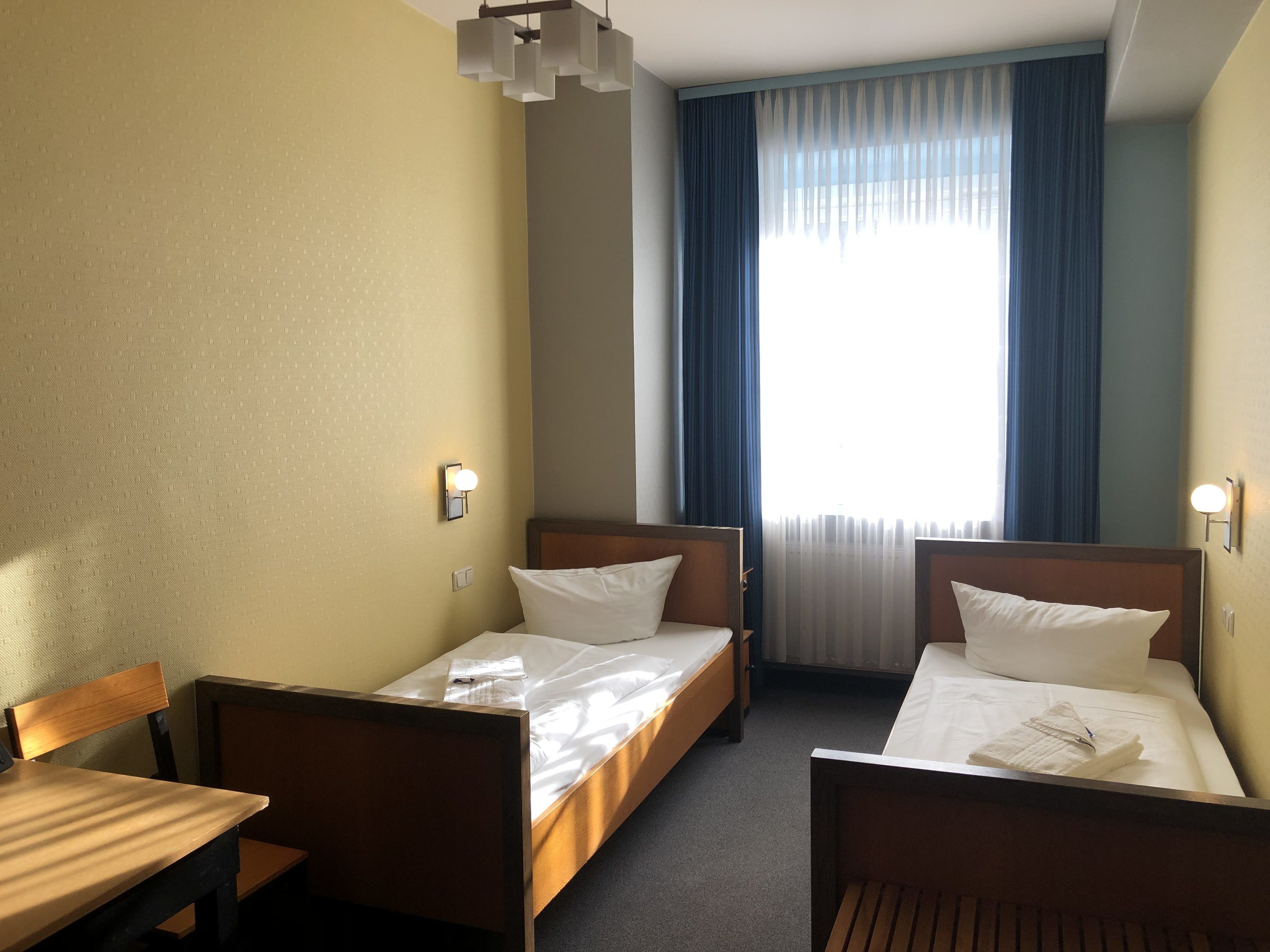A hotel room with two single beds, a wooden table, a chair, and a window with blue curtains.
