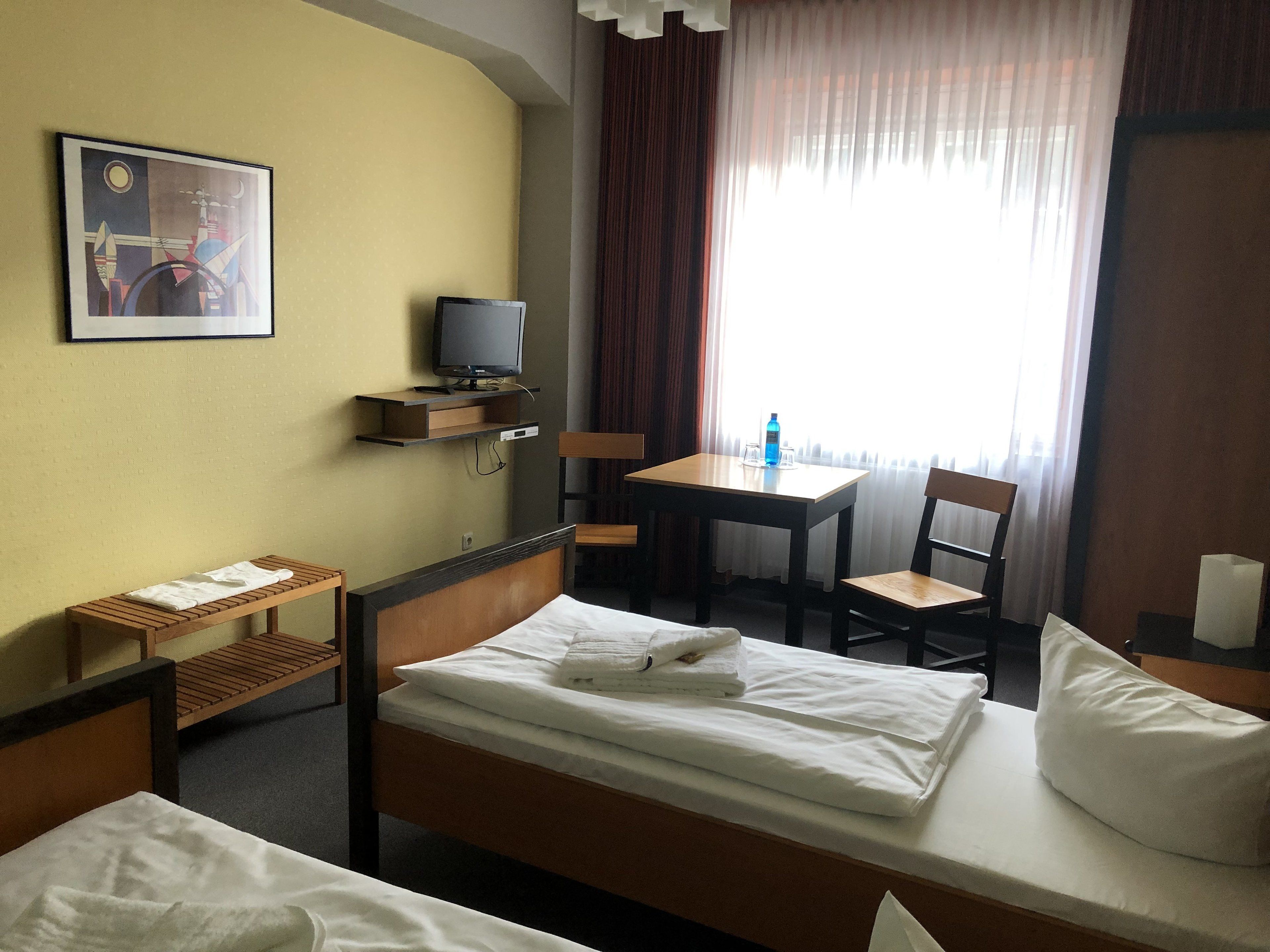 Hotellihuone, jossa on sänky, työpöytä tuolilla, hyllyllä oleva televisio ja ikkuna, jossa on verhot.