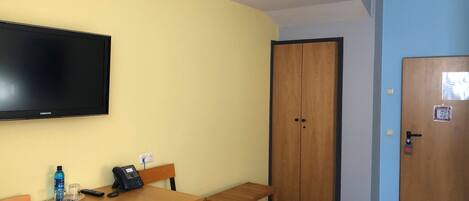 A room with a flat-screen TV, a wooden table with chairs, a small wooden stool, a telephone, and a door with a lock and a QR code.