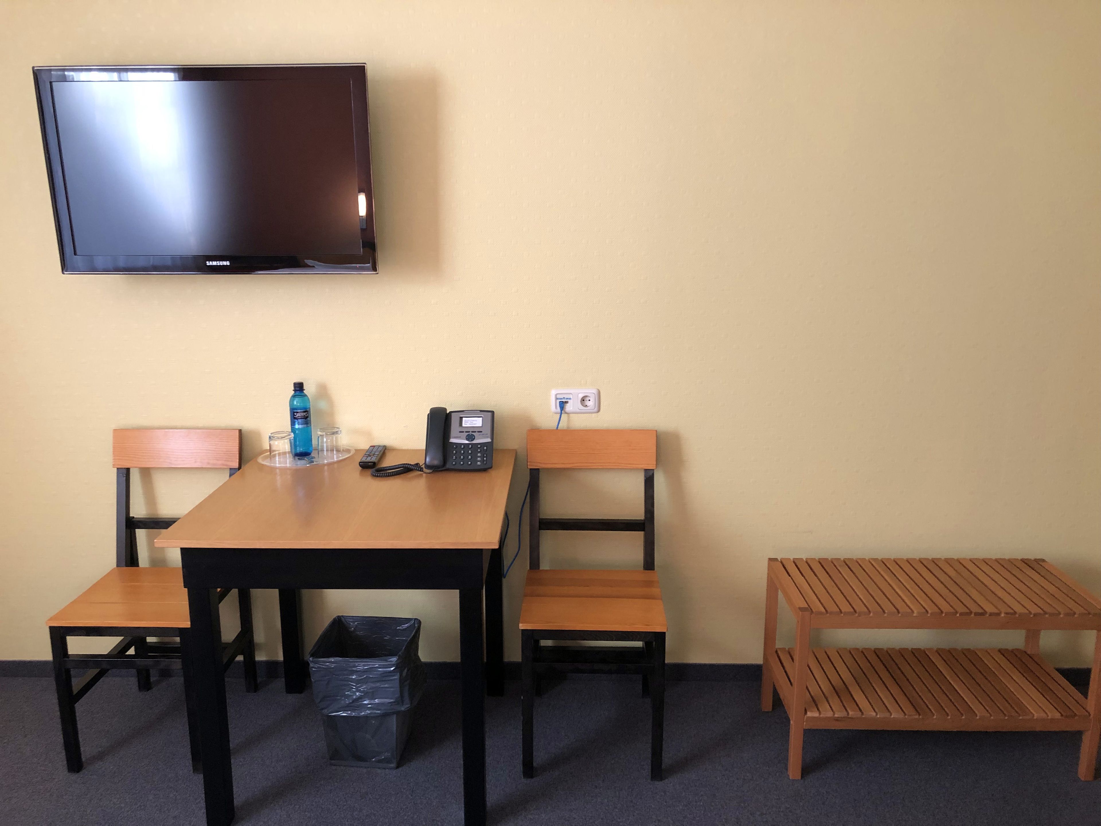 A hotel room with a wooden table, two chairs, a television, a telephone, a trash bin, and a small wooden bench.