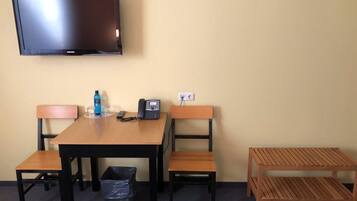 A hotel room with a wooden table, two chairs, a television, a telephone, a trash bin, and a small wooden bench.