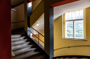 Uma escadaria com corrimão, uma janela com vista para vegetação e uma parede com esquema de cores amarelo e azul.