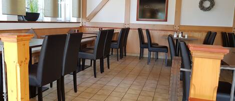 Interior de restaurante con mesas y sillas de madera, una pizarra con escritura y una planta en maceta junto a la ventana.