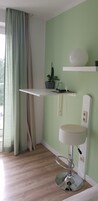 Una habitación de esquina con una pared verde claro, un estante blanco con una planta en maceta, un lavamanos pedestal blanco y suelo de madera.