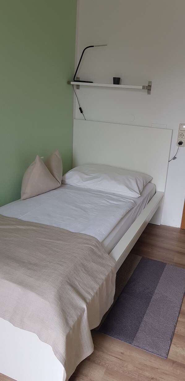 Una cama bien hecha con una manta color beige, un cabecero blanco y una pequeña estantería arriba.