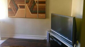 A living room with a flat-screen TV on a stand, a wall-mounted abstract art piece, and a wooden floor. - Geomara 2 Bdroom Ppl Center (Nairobi)