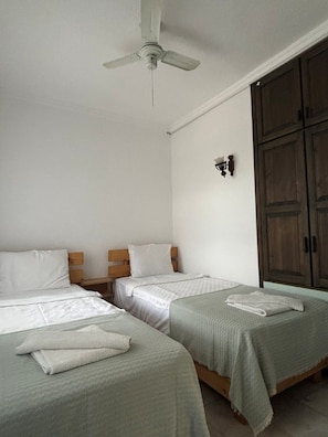 Una habitación con dos camas individuales, un ventilador de techo, un armario de madera y una lámpara fijada a la pared.