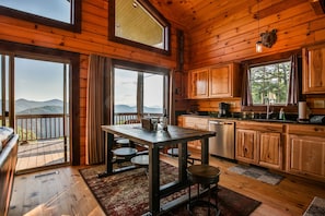 Una cabaña de madera con comedor, cocina y grandes ventanales que ofrecen vistas a la montaña.