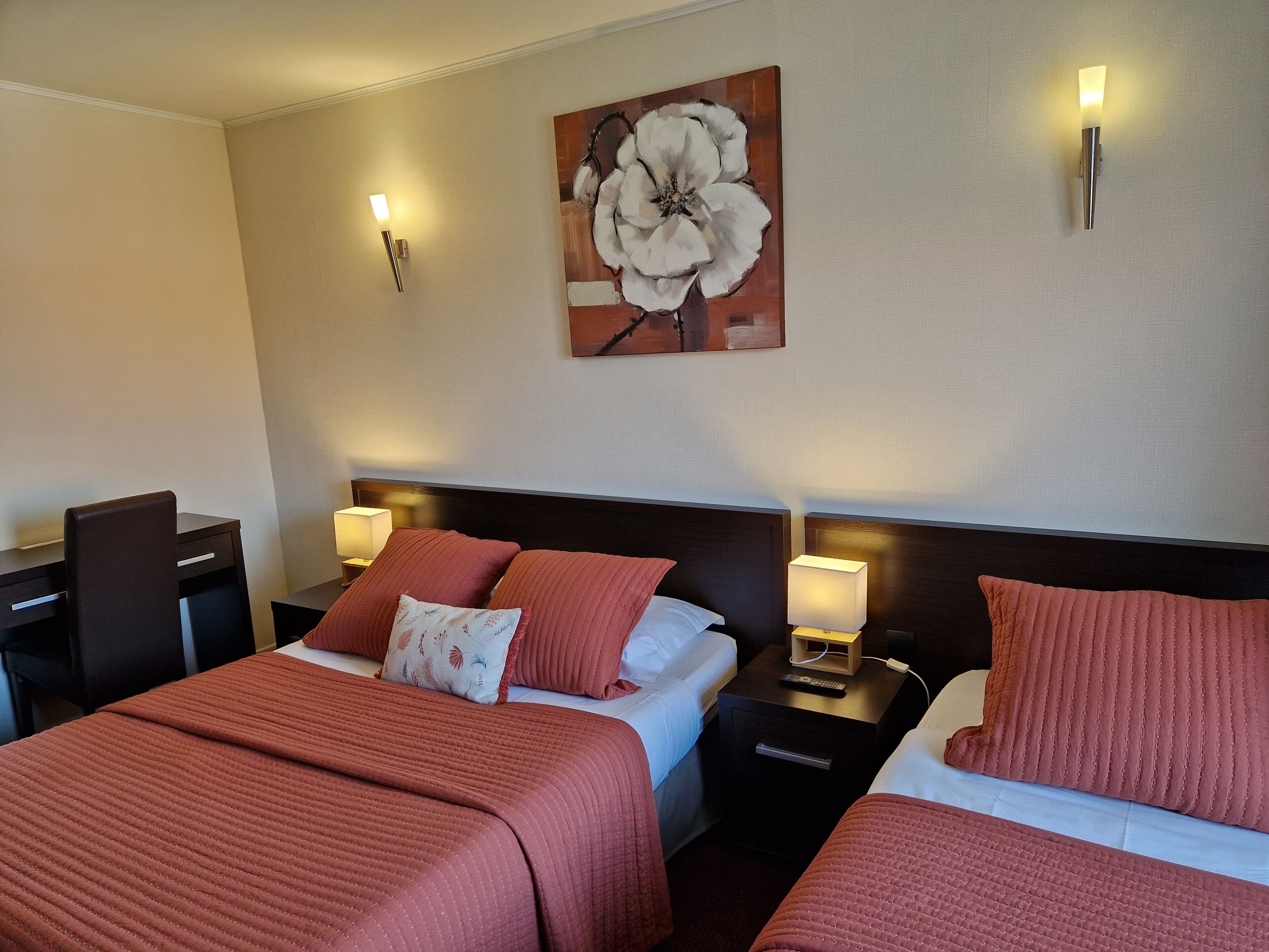 Ein Hotelzimmer mit zwei Betten, jeweils mit roter Bettdecke und passenden Kissen. Es gibt Nachttische mit Lampen, ein großes Blumenbild an der Wand und einen Schreibtisch mit Stuhl.