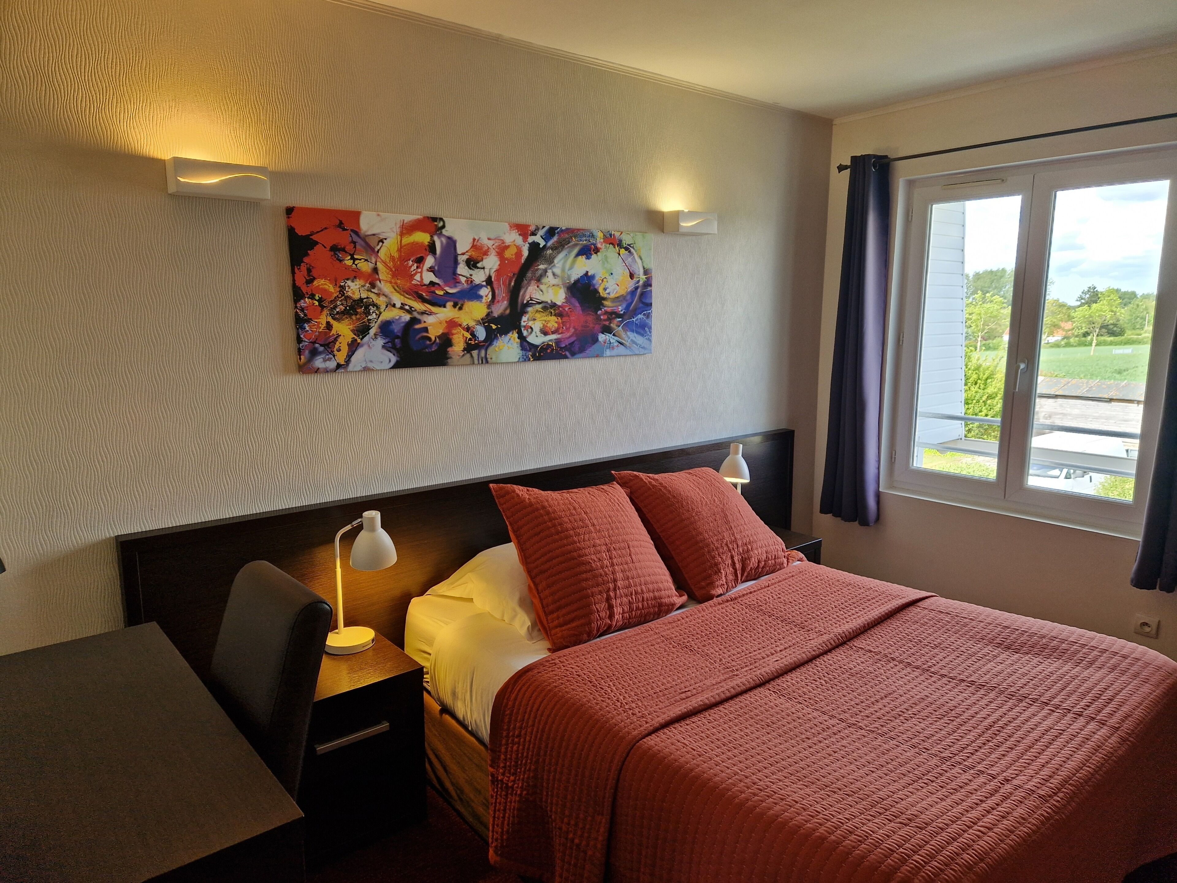 A hotel room with a bed, a desk, a lamp, a painting on the wall, and a window with curtains.