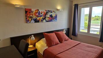 A hotel room with a bed, a desk, a lamp, a painting on the wall, and a window with curtains.