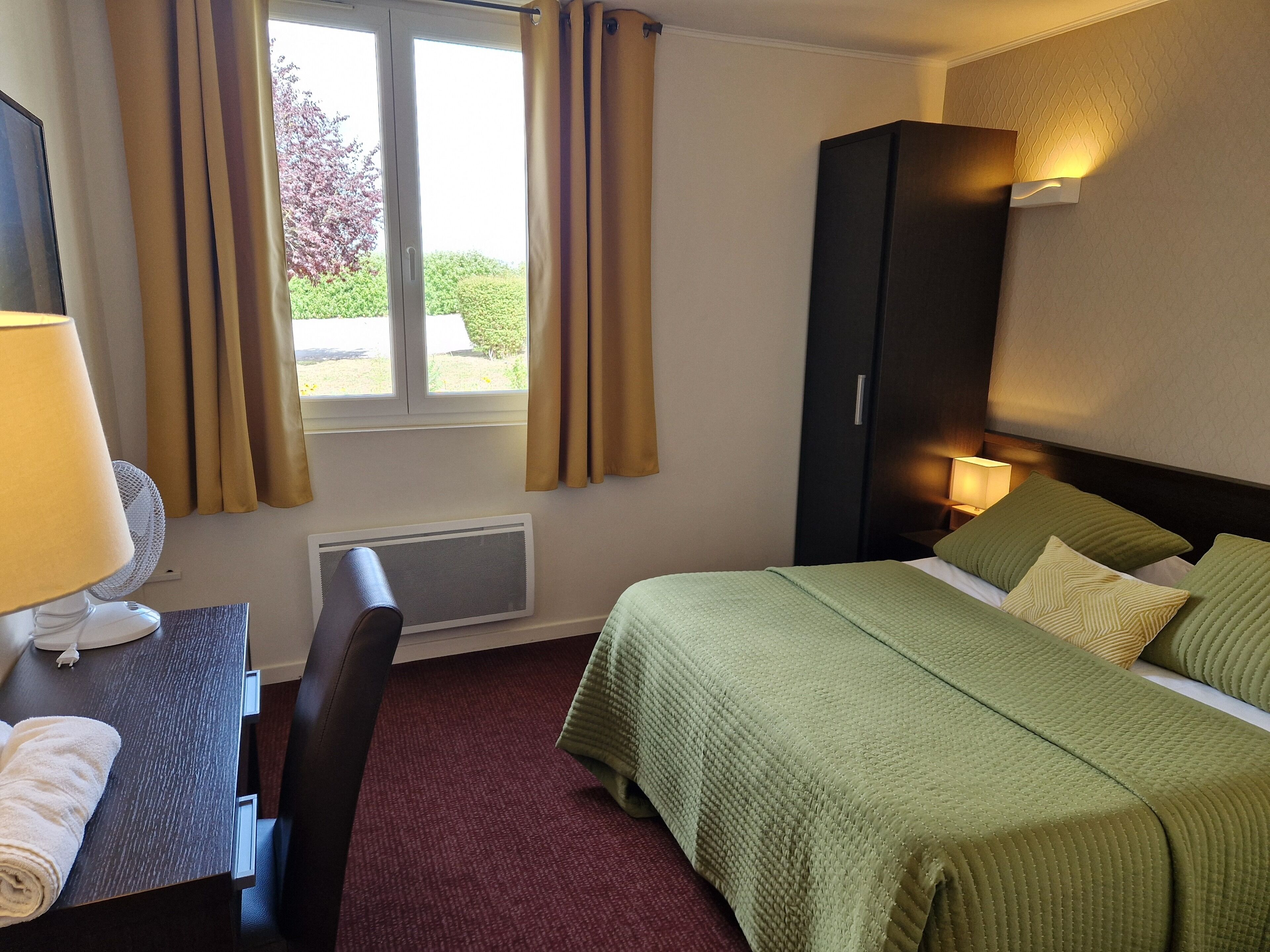 A hotel room with a bed, a desk with a lamp, a chair, a wardrobe, and a window with curtains.