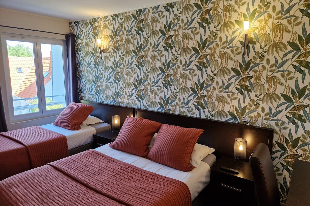 A hotel room with two beds, a large window, and a wall with a leafy pattern.