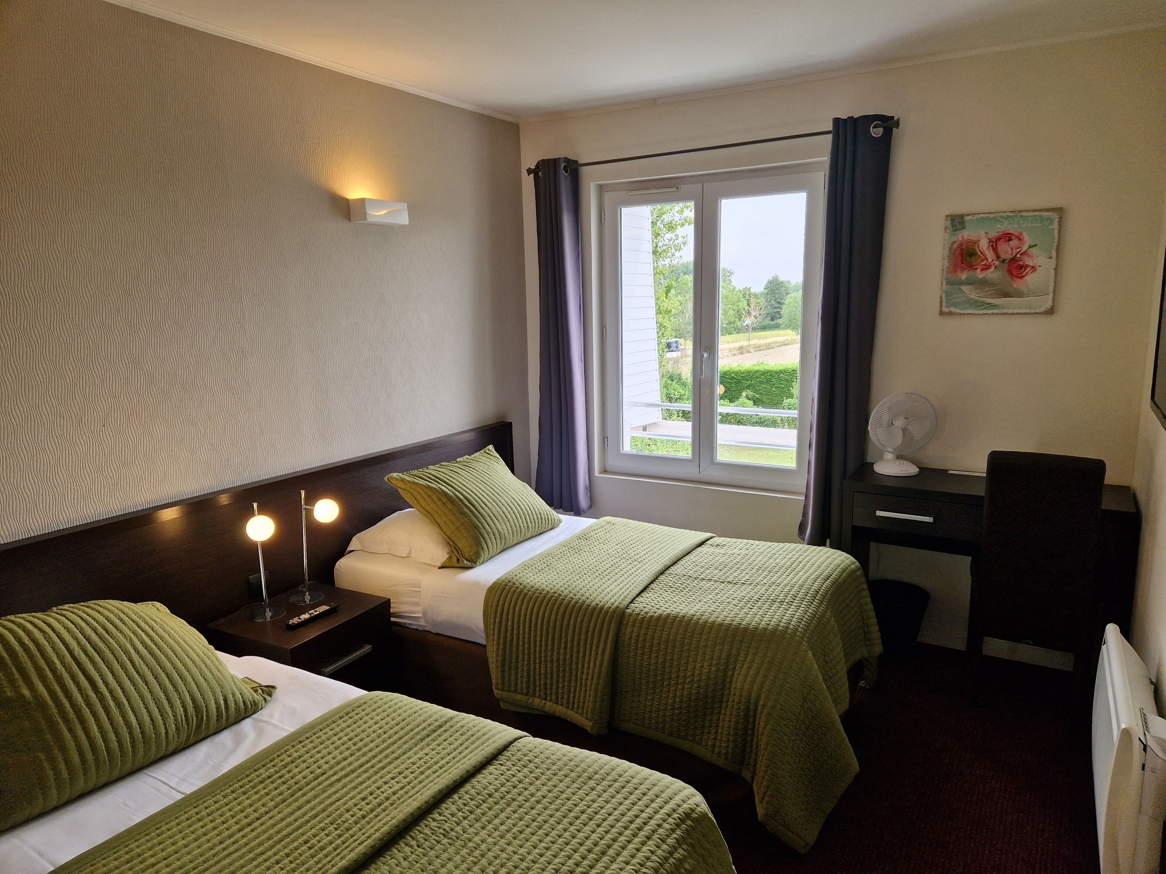 A hotel room with two beds, a desk, a chair, a window with curtains, a wall-mounted light, and a picture of roses.