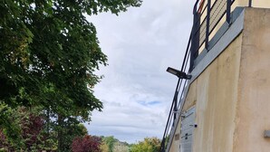 Un chemin bordé d’une rambarde métallique, qui mène à un bâtiment à la façade beige et à l’escalier extérieur en métal noir.